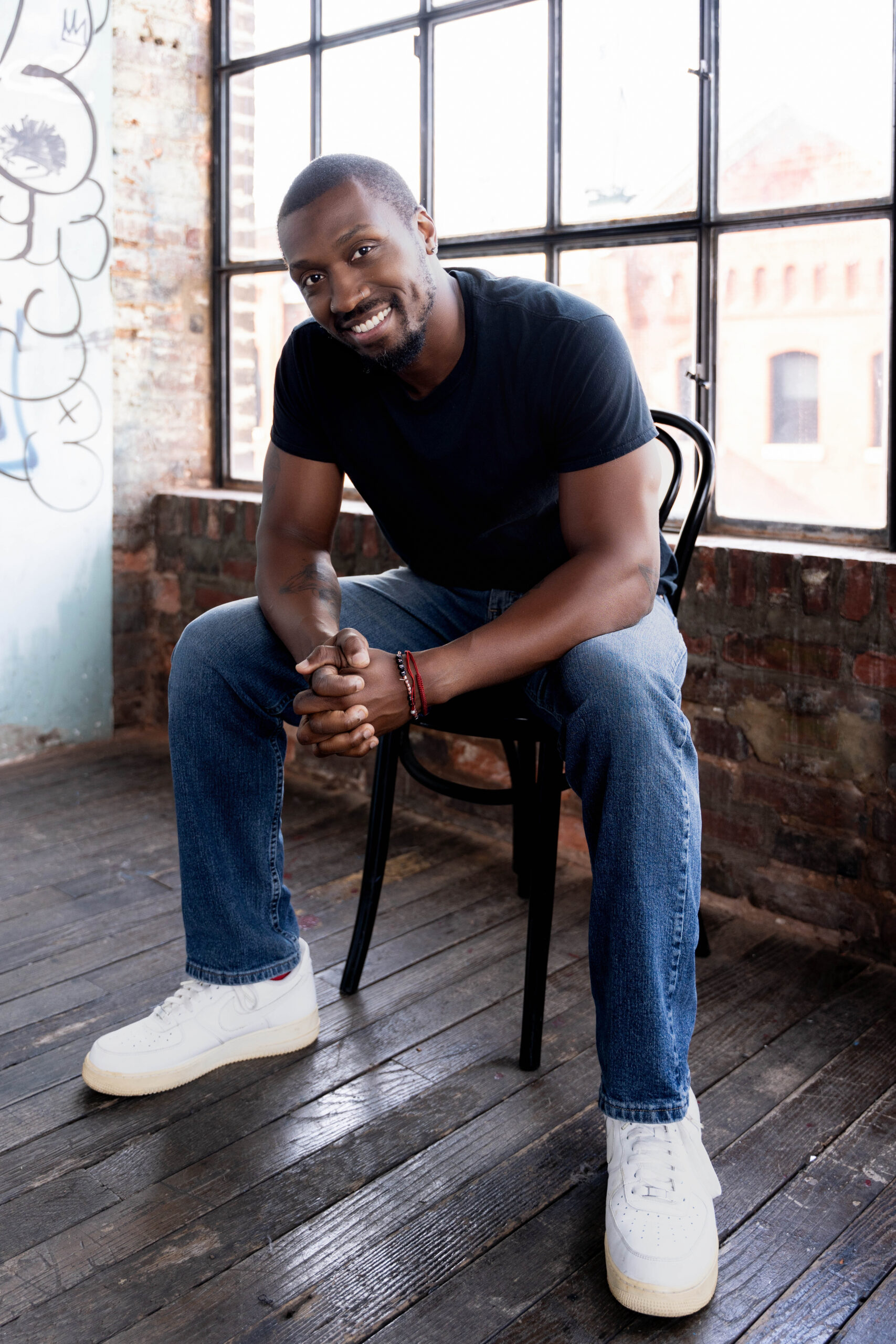 A man in jeans and black shirt sits in a black chair under a window