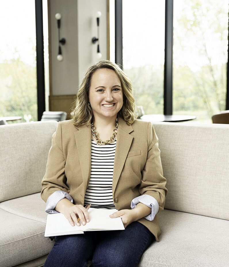 A smiling woman in a brown blazer sits on a couch in a lobby writing in a notebook during Baltimore Personal Brand Photos