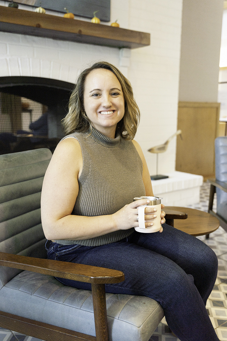 A smiling woman sits in a cafe chair holding a cup of coffee in jeans