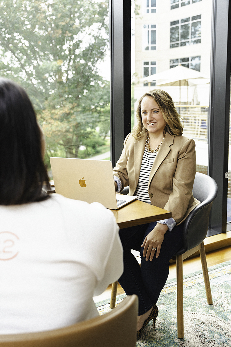 A woman chats with a client at a wooden office table in a window with her laptop during a DC personal branding session