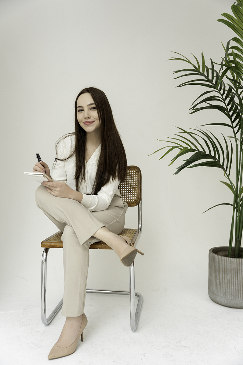 A woman sits in a wooden chair taking notes in tan pants and white blouse