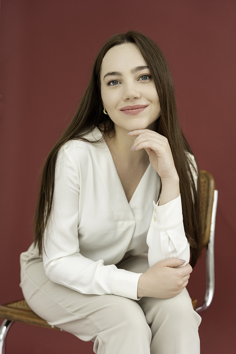 A woman in a white blouse sits on a wooden chair in a red studio for Personal Branding Photos