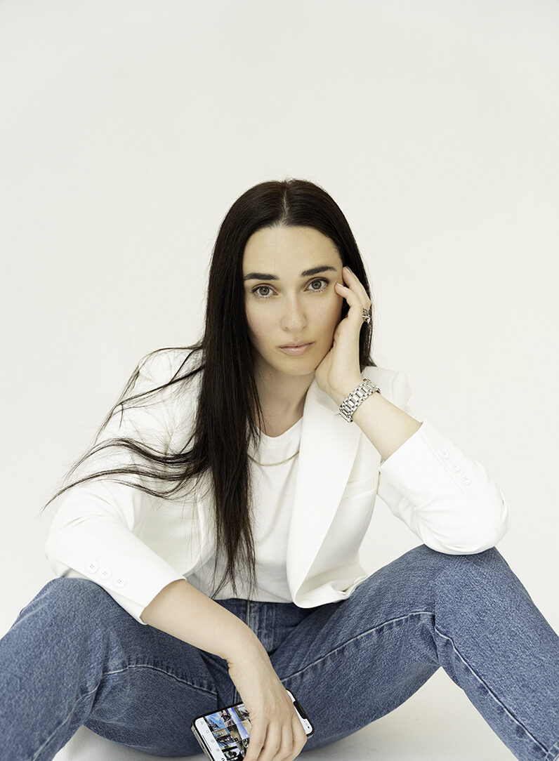 A woman sits on the floor of a studio holding her phone in jeans and white blazer