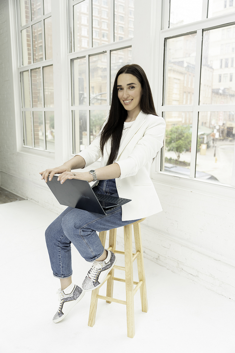 A woman sits on a wooden stool in a studio window with a laptop in her lap