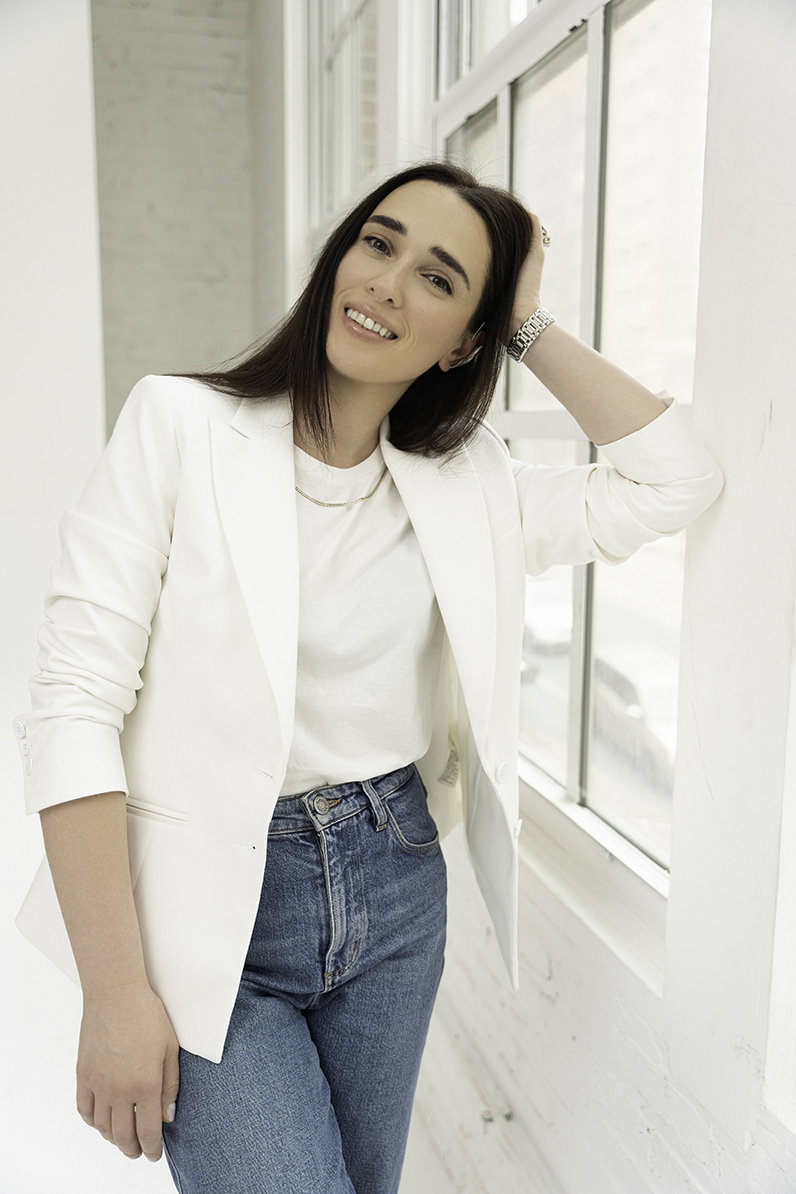 A woman in a white blazer and jeans leans in a window with a hand in her hair to Update Personal Brand Photos