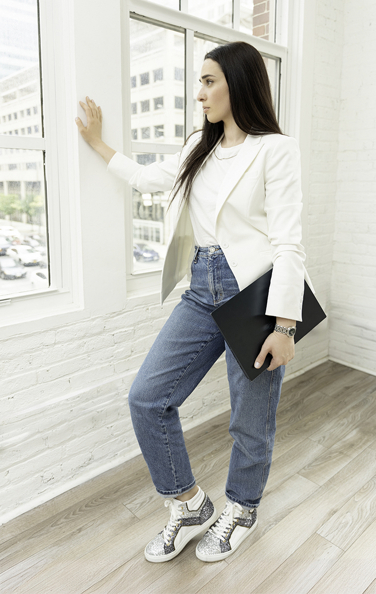 A woman in jeans holds a notebook leans against a window gazing out it to Update Personal Brand Photos